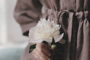 Stylish woman in linen dress holding white peony in sunny light. Slow life. Young female in boho rustic dress with white peony. Aesthetic moment