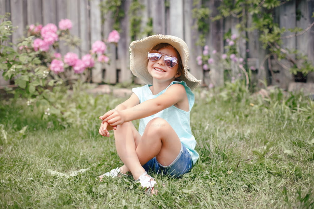 Porträt eines niedlichen entzückenden Kindermädchens mit Sonnenbrille und Strohhut, das draußen auf Gras sitzt. Glücklich lächelndes kaukasisches Kind, das Spaß im heimischen Hinterhof hat. Erstaunliche fröhliche Sommer- und Lifestyle-Kindheit.