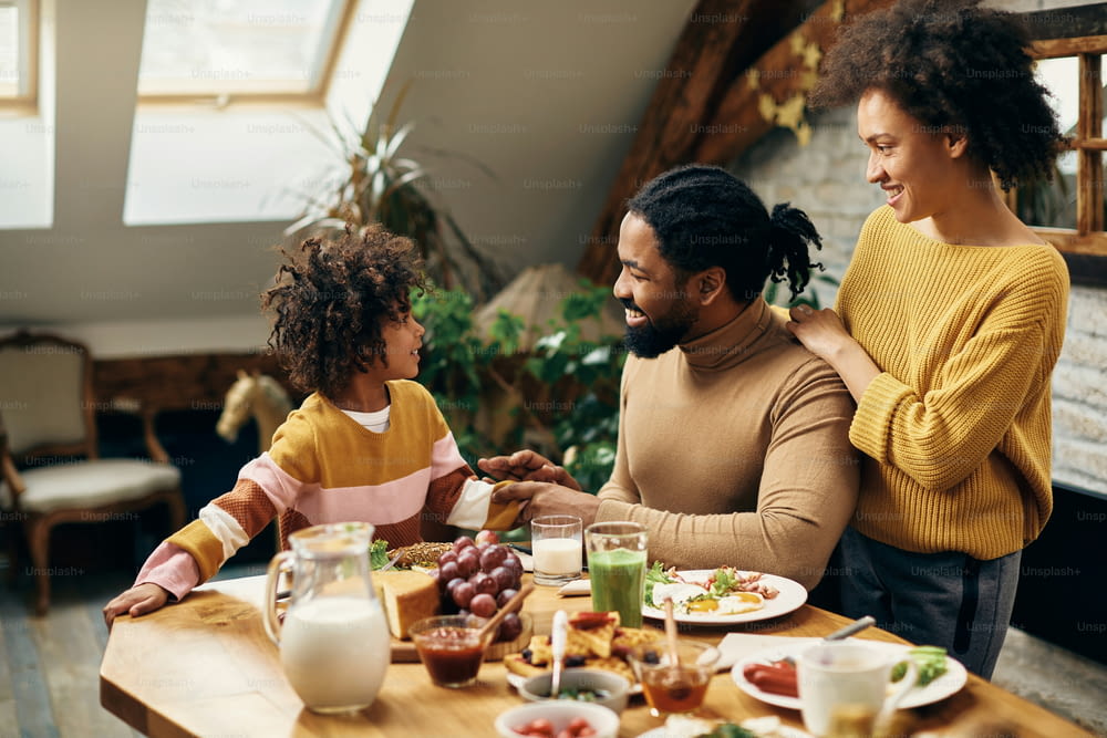 Happy black parents and their small daughter communicating during a meal in dining room.