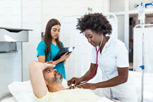 의사가 맥박을 확인하는 동안 침대에 누워 있는 입원 남자. 의사와 간호사가 병실에서 노인 남성 환자를 검사하고 있다.