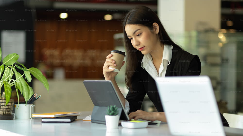 Portrait of businesswoman holding paper cup while working with digital tablet in office room