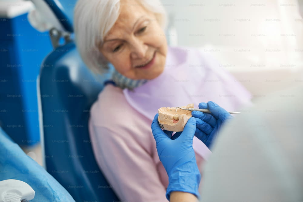 Dental surgeon using periodontal probe for pointing a sick tooth on a lower jaw mold for senior lady