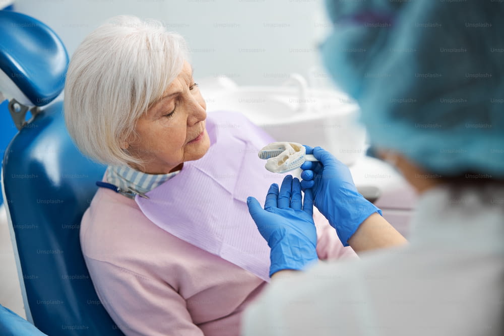 수술의자에 앉은 은퇴한 여성에게 실리콘에서 탄력 있는 치아 인상을 제공하는 치과 의사