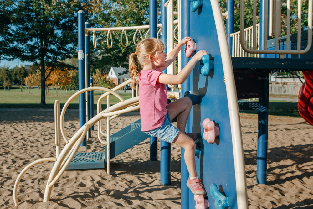 Little preschool girl climbing rock wall at playground outside on summer day. Happy childhood lifestyle concept. Seasonal outdoor activity for kids. Strong girl female power.
