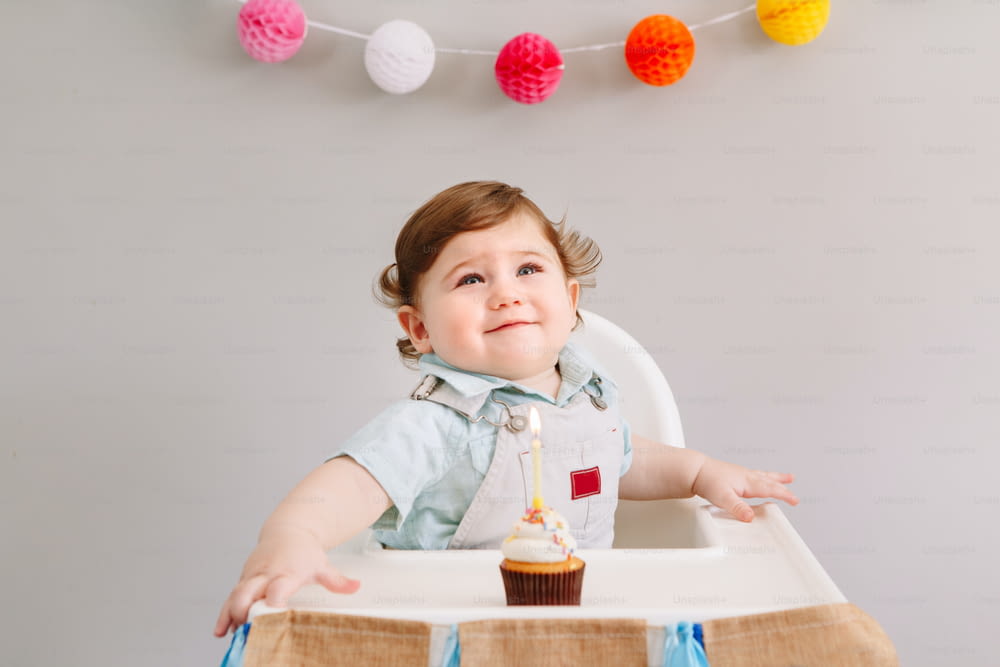 Happy smiling cute Caucasian baby boy celebrating his first birthday at home. Child kid toddler sitting in high chair eating tasty cupcake dessert with lit candle. Happy birthday concept.