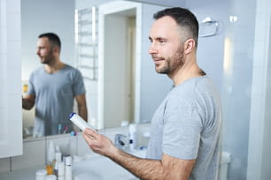 Smiling gentleman with cosmetic product in his hand standing in front if the mirror in bathroom