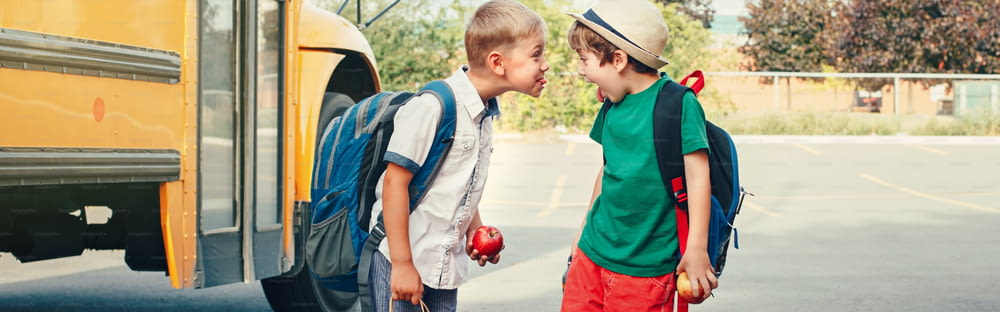 Two funny happy Caucasian boys kids showing tongues each other near yellow bus on 1 September day. Education back to school. Children learn and study. Web banner header for website.