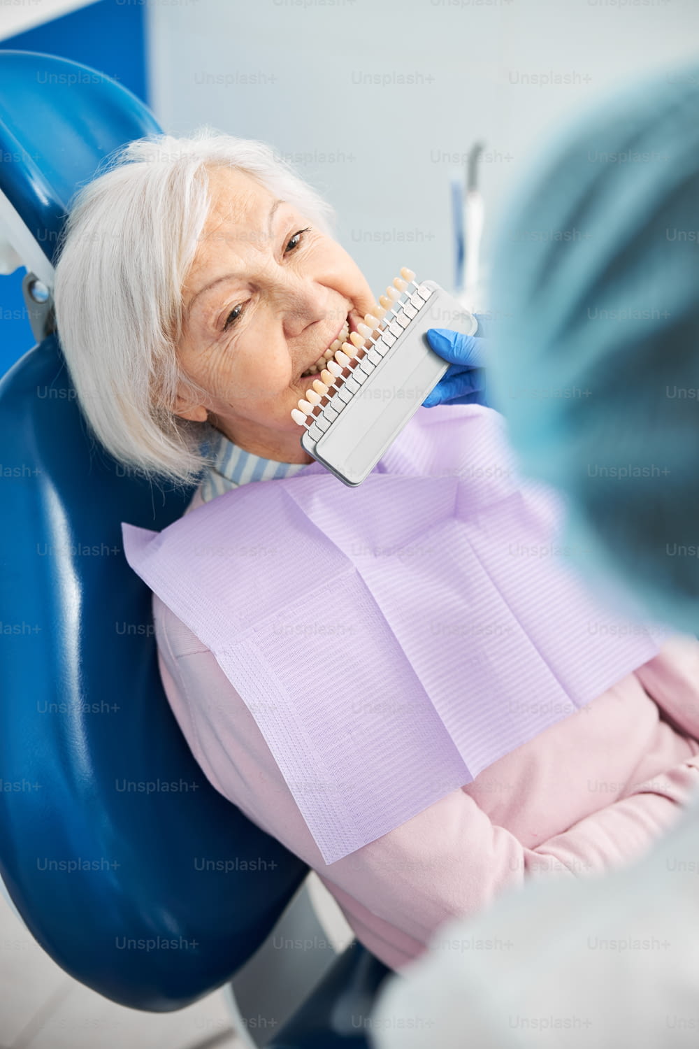Retired woman in dentist armchair showing teeth while dental practitioner holding dental shade chart to her mouth