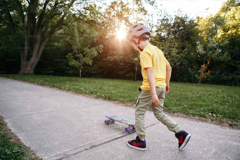 회색 헬멧을 쓴 소년이 여름날 공원의 도로에서 스케이트보드를 타고 있다. 계절에 따라 야외 어린이 활동 스포츠. 건강한 어린 시절의 생활 방식. 스케이트 보드를 타는 법을 배우는 소년. 뒤에서 본 모습.