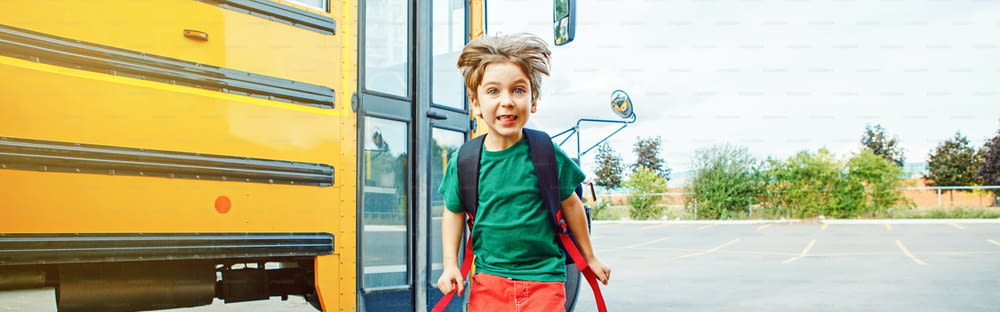 Emotional Caucasian boy student kid with funny face expression jumping near yellow bus on 1 September day. Education back to school. Child ready to learn, study. Web banner header for website.