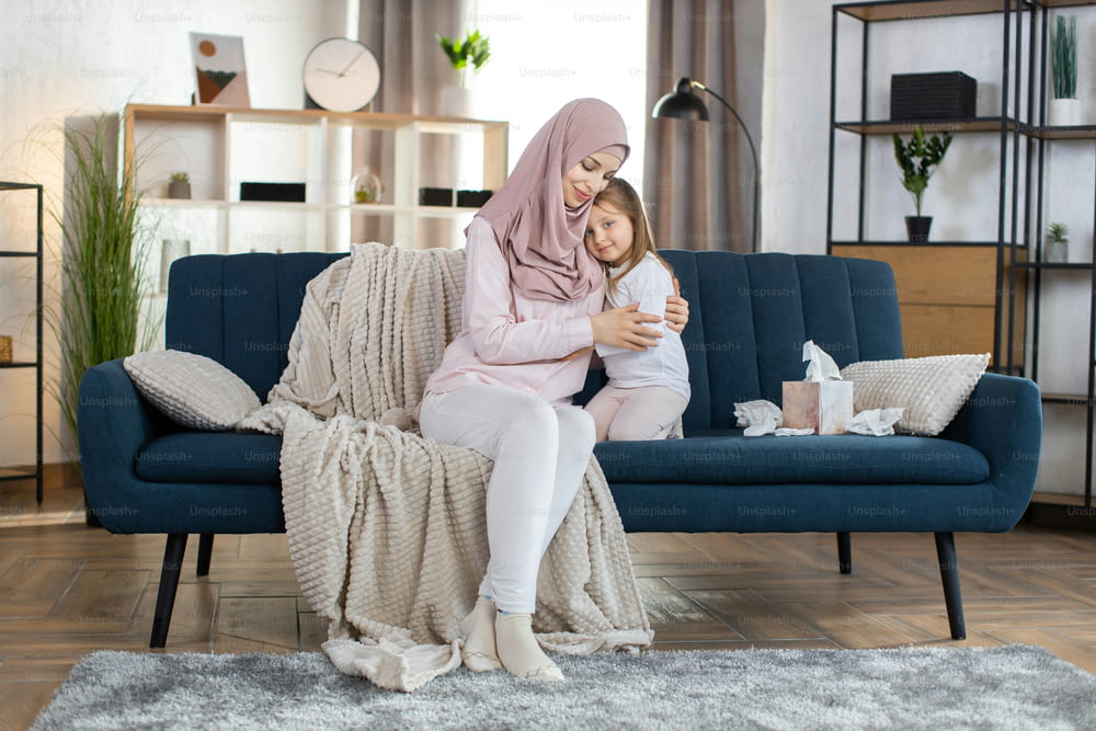 Happy motherhood, Mother day concept. Pretty young smiling Muslim woman mother hugging cute little daughter, posing to camera at home in cozy living room. Mom and child in domestic wear