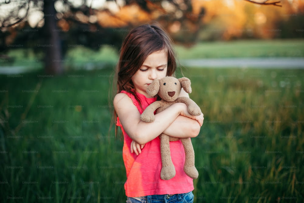 Triste ragazza caucasica sconvolta che abbraccia il giocattolo. Il bambino abbraccia l'orso morbido di peluche nel parco all'aperto. Bambino solitario perso fuori. Stile di vita infelice, problemi infantili.
