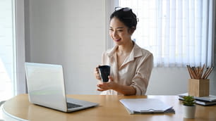 Femme d’affaires joyeuse tenant une tasse de café et utilisant un ordinateur portable passant un appel vidéo avec des collègues.
