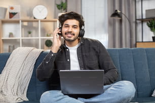 Giovane uomo arabo o indiano di bell'aspetto, seduto sul divano di casa, che lavora su un pc portatile e ascolta la sua musica preferita allo stesso tempo utilizzando gli auricolari.