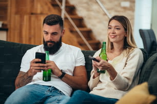 Giovane coppia che si rilassa a casa e beve birra mentre utilizza uno smartphone