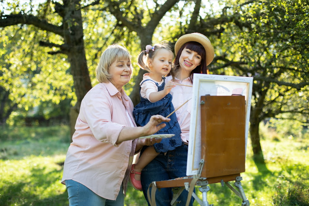 Lovely Caucasian three generation family, grandmother, mother and little cute child kid girl, standing outside in summer spring park and painting a picture on wooden easel.
