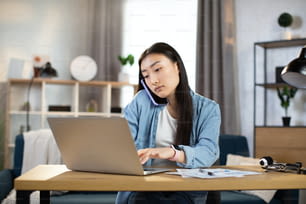 Giovane donna asiatica in abito casual che parla al telefono cellulare e digita sul laptop mentre è seduta sul posto di lavoro. Concetto di comunicazione aziendale, freelance, lavoro online.