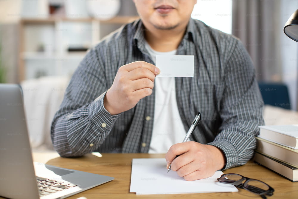 Close up of young man win casual outfit sitting at desk with wireless laptop and showing empty white card. Space for text on business card.
