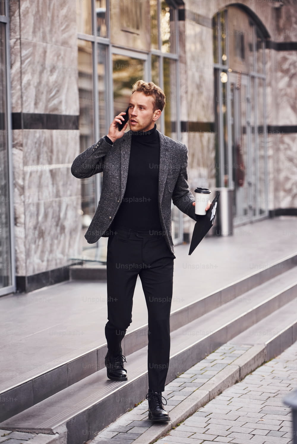 Homem em trajes formais elegantes com copo de bebida e bloco de notas nas mãos está do lado de fora contra o edifício moderno ter uma conversa ao telefone.