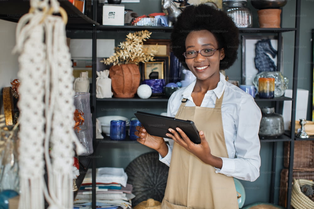 Close up of smiling pretty afro american woman in eyegasses using digital tablet for making inventory at decor shop. Attractive saleswoman in apron standing with modern gadget in hands.