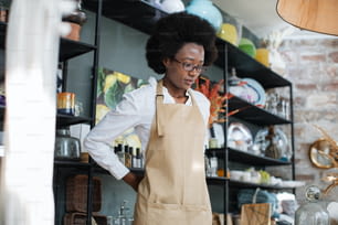 Atractiva mujer afroamericana con delantal beige durante el proceso de trabajo en la tienda de decoración. Variedad de cosas exclusivas para la decoración.