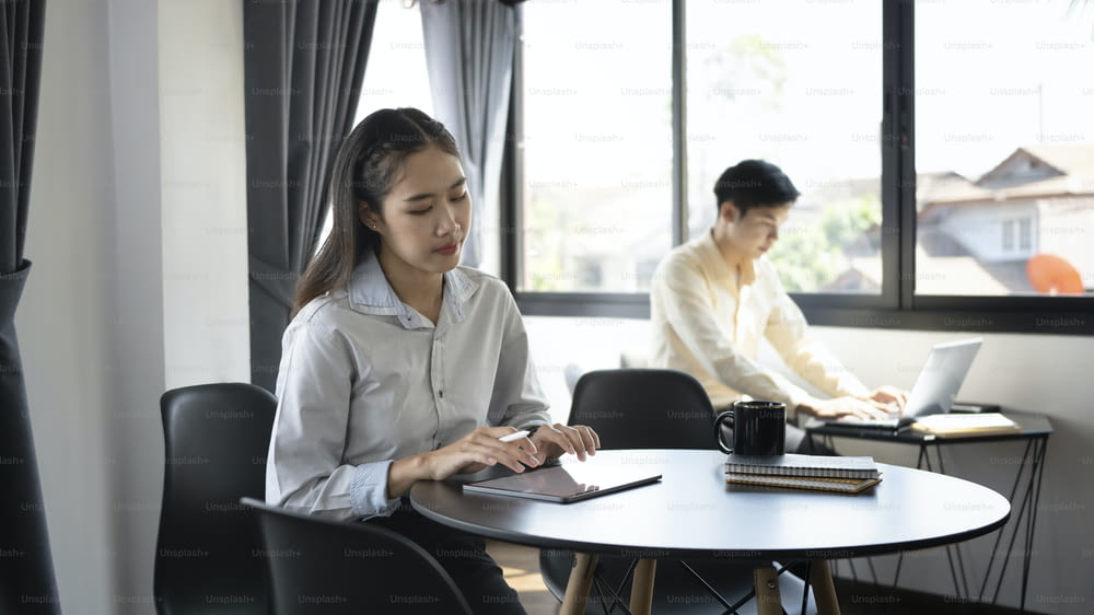 Young asian office worker using digital in office and her colleague sit-in in background.