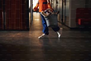 Foto recortada de una mujer con uniforme y guantes de goma cargando un cubo de plástico con suministros de limpieza
