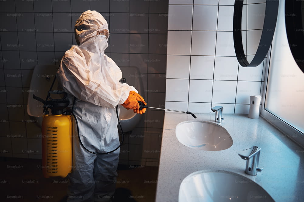 Side view of a woman in safety goggles and in rubber gloves sanitizing the sink in the public bathroom