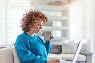 Smiling young redhead holding a cup of coffee while working on her laptop in her home. Happy Girl Relaxing on Comfortable Couch and Using Laptop at Home, Surfing Internet