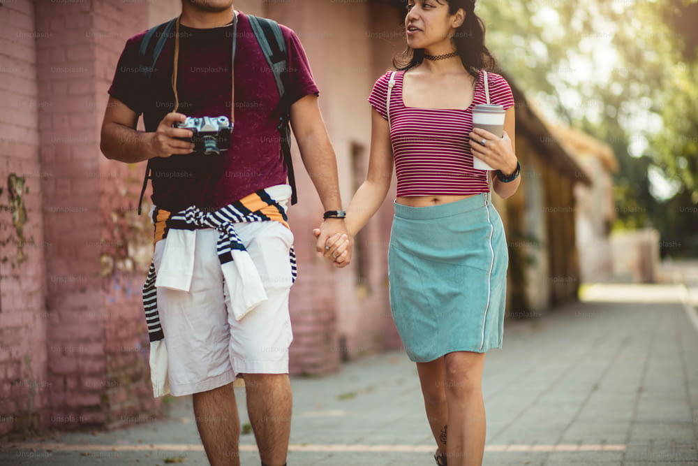 Young couple walking trough street and holding hands.  Man with camera and girl with cup of coffee.