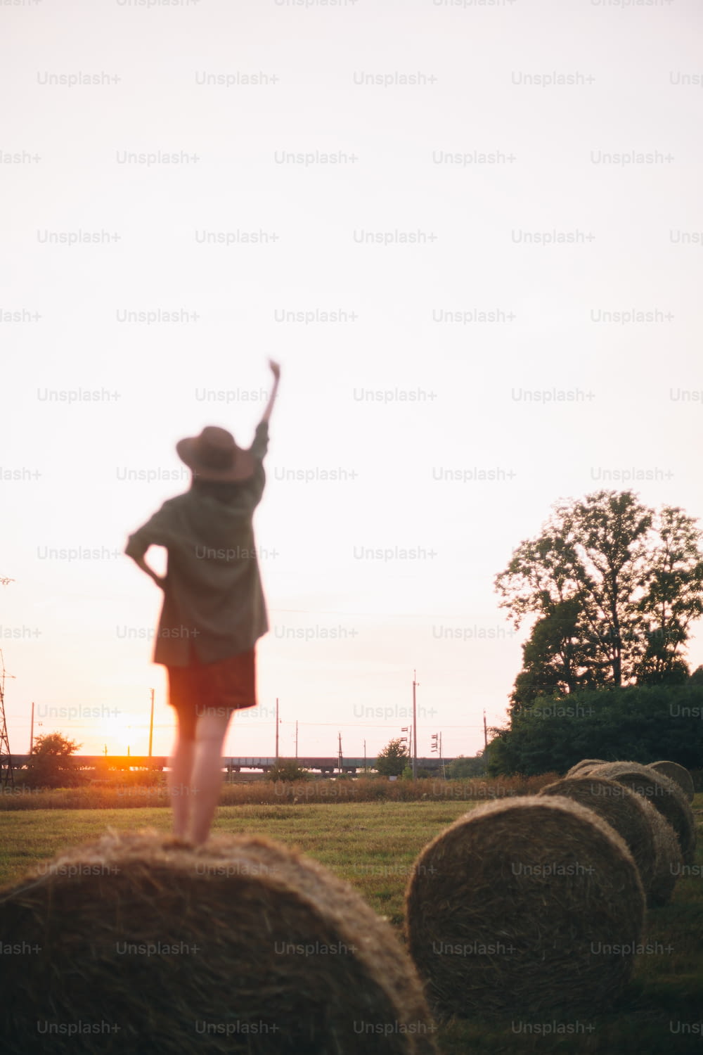 Unscharfe Silhouette einer glücklichen Frau auf Heuhaufen, die den Abend genießt und sich darauf konzentriert, im Sommerfeld bei Sonnenuntergang zu trainieren. Junges, glückliches Weibchen entspannt sich auf Heuballen auf dem Land. Atmosphärischer Moment
