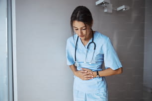 Nette junge medizinische Mitarbeiterin, die an der Wand steht und die Hände auf den Bauch legt, während sie Bauchschmerzen in der Klinik hat
