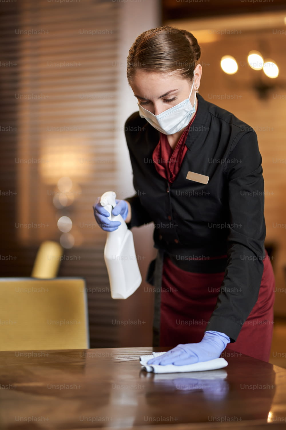 Attentive waitress wearing face mask and holding spray bottle with disinfectant while wiping the table