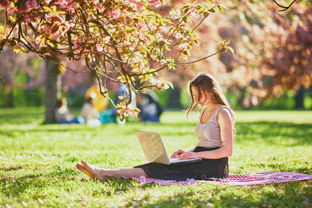 Beautiful young girl working on her laptop in park during cherry blossom season. Young woman in famous Park of Sceaux near Paris, France. Freelance, distance learning or remote work concept