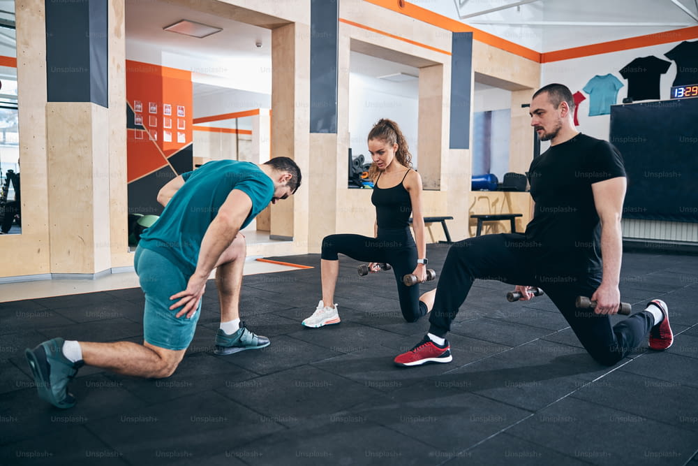 Focused woman and serious man performing the low lunge with hand weights assisted by their fitness coach
