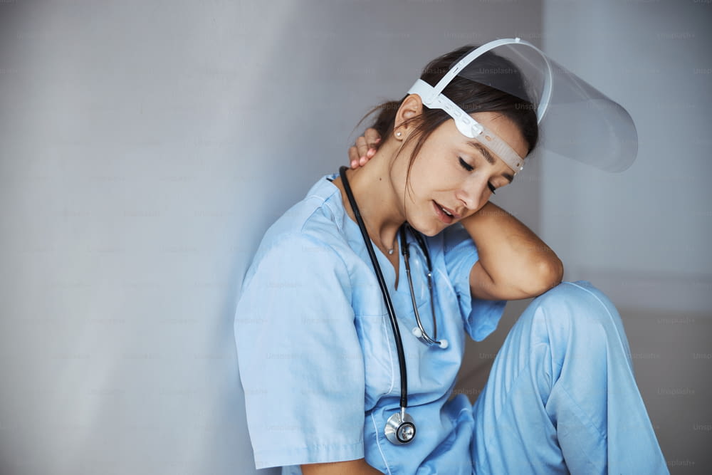 Giovane operatrice medica stanca che indossa una maschera protettiva per il viso e un'uniforme ospedaliera mentre soffre di affaticamento