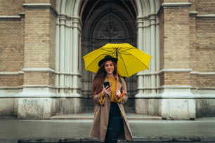 Mujer joven hermosa que usa un teléfono inteligente y sostiene un paraguas amarillo mientras camina en una ciudad en un día lluvioso