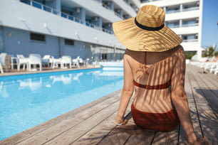 夏休みを楽しんでいるプールの木製の桟橋のヤシの葉の影の下でリラックスする帽子をかぶった美しい女性。トロピカルホテルリゾートのプールで日焼けするスリムな若い女性