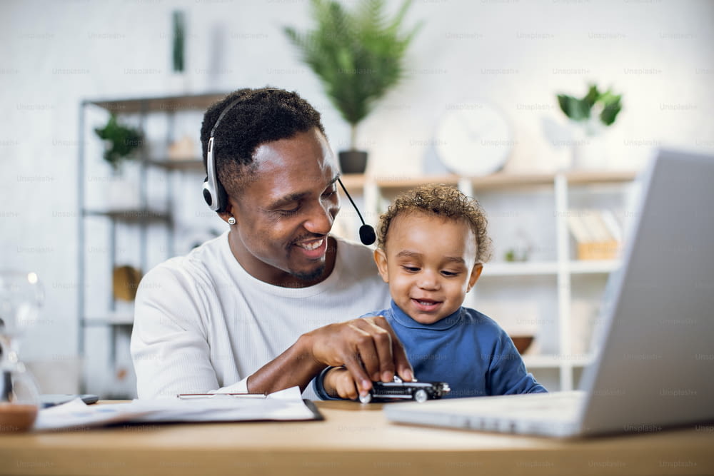Père afro-américain dans un casque jouant avec un fils mignon et menant une réunion en ligne sur un ordinateur portable moderne. Jeune homme assis à la maison avec un petit garçon et travaillant à distance.