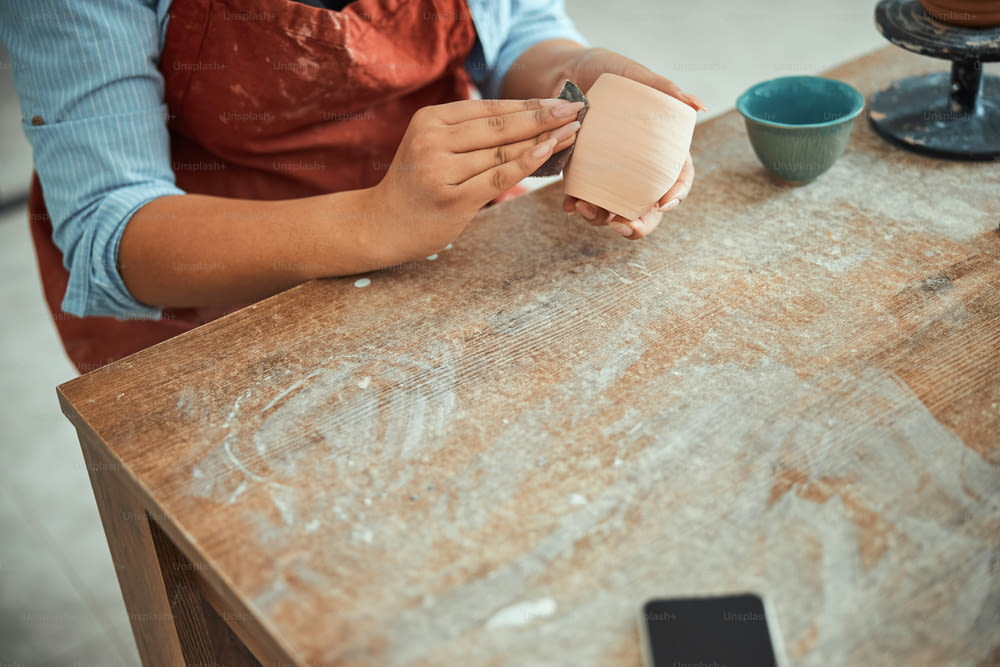 Close up of young woman potter polishing and smoothing earthenware with sandpaper while sitting at the table in pottery studio