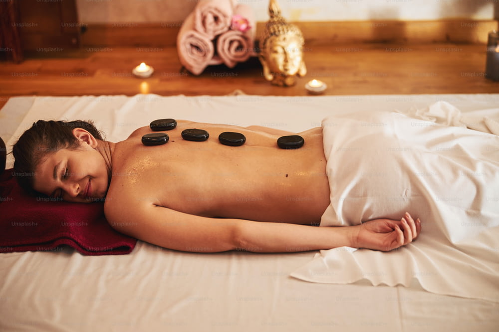 Mujer acostada con comodidad en un colchón blanco mientras tiene piedras de masaje distribuidas en su espalda