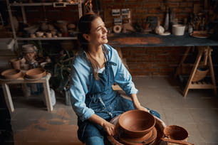 Positive craft woman wearing apron sitting near the pottery wheel while looking away during the process manufacturing of ceramic bowl in art studio