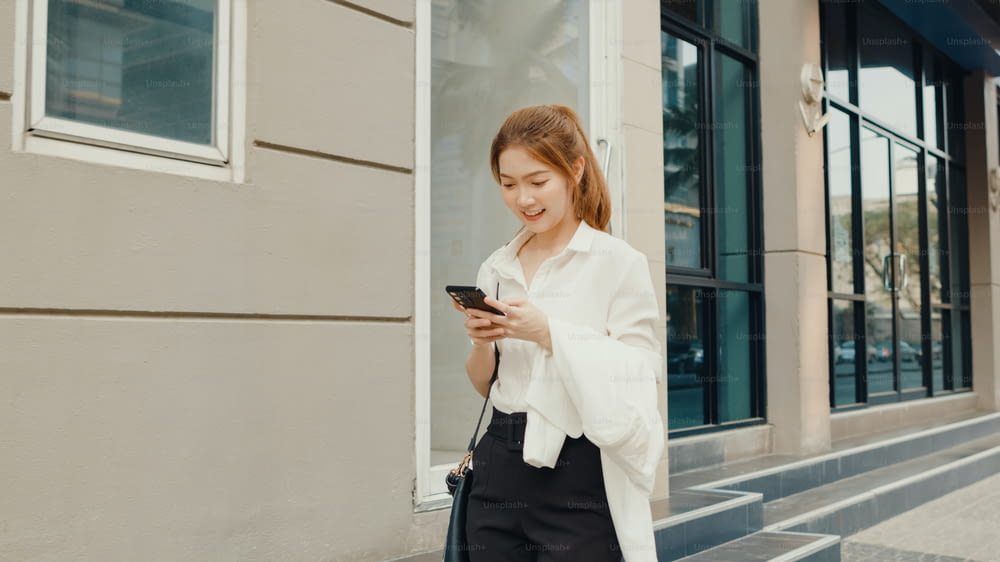 Successful young Asia businesswoman in fashion office clothes using smart phone and typing text message while walking alone outdoors in urban modern city in the morning. Business on the go concept.
