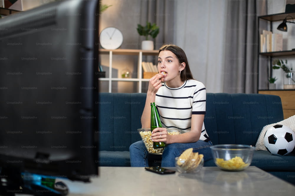 Bela jovem mulher em traje casual comendo lanches e bebendo cerveja enquanto assiste a jogo de futebol na TV em casa. Conceito de torcedor, lazer e esporte.