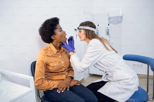 Doctora otorrinolaringóloga examinando la boca del paciente en una clínica moderna. Médico en el hospital revisando el dolor de garganta y las amígdalas de una joven paciente afroamericana