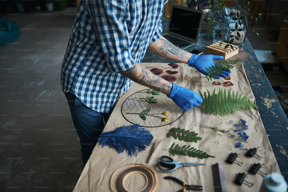 Close up of young man with tattoos on arms holding plants while standing by the table with herbarium