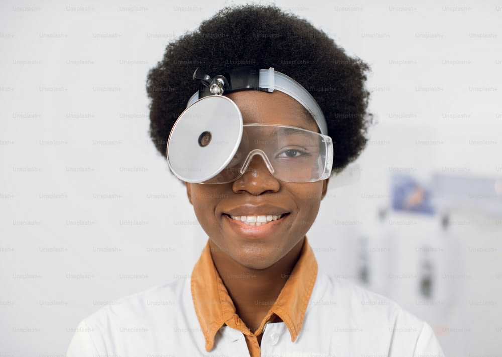 Gros plan d’une jeune femme afro-américaine souriante et amicale, médecin et oto-rhino-laryngologiste, portant un réflecteur frontal et des lunettes de protection, posant devant la caméra dans son bureau moderne