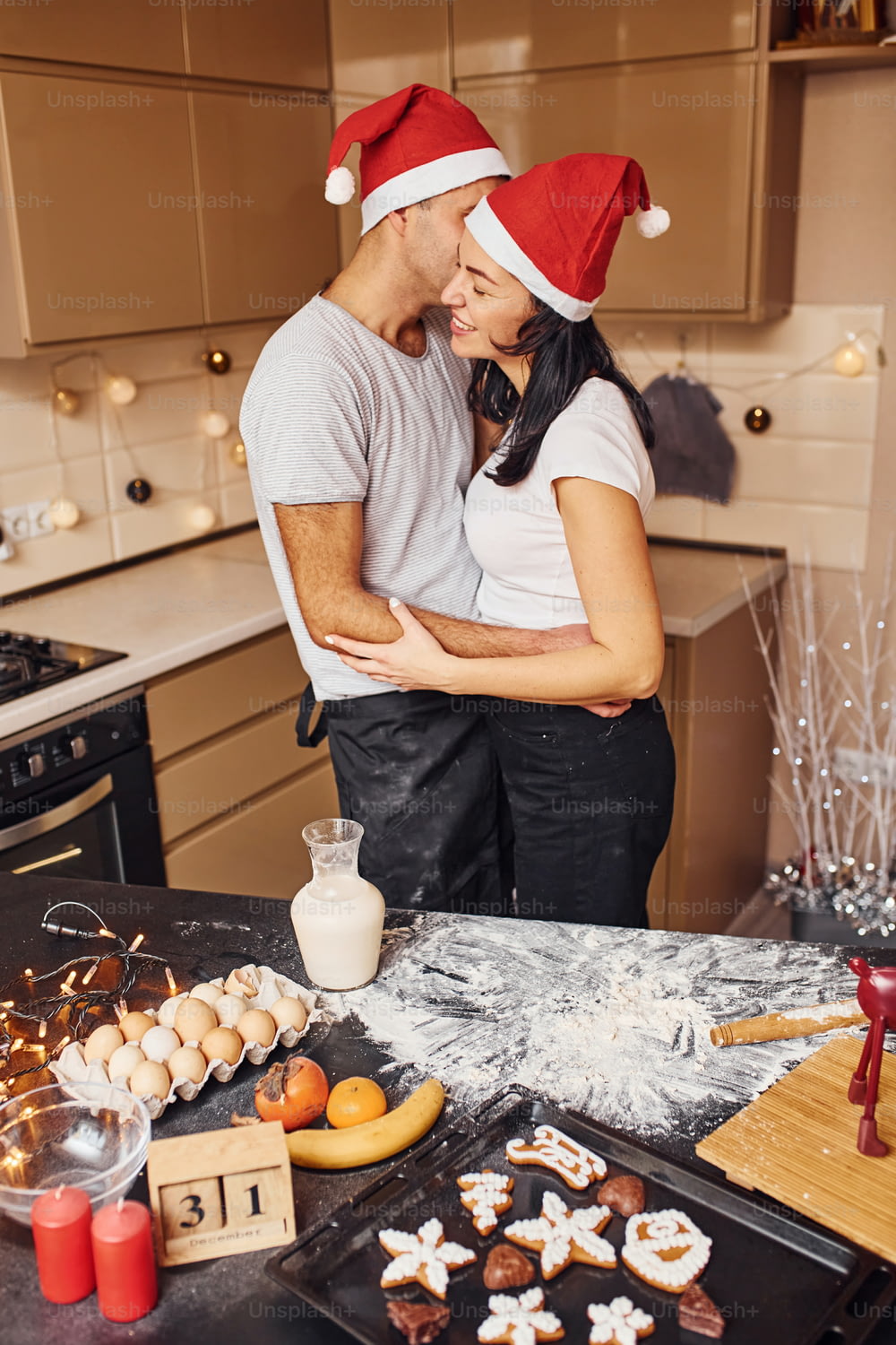 Embracing and kissing. Couple with santa hats standing on the kitchen together indoors.