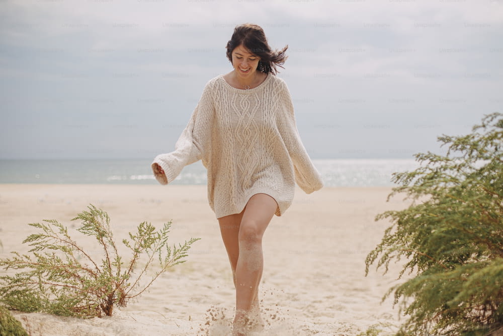 Hermosa mujer feliz con cabello ventoso caminando en la playa de arena en el fondo de la hierba verde y el mar, momento tranquilo y tranquilo. Mujer joven con estilo en suéter de punto beige que se relaja en la costa fría