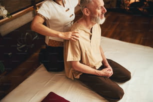 Vista dall'alto di un maschio brizzolato rilassato seduto in ginocchio mentre una donna professionale sta trattando manualmente le sue spalle
